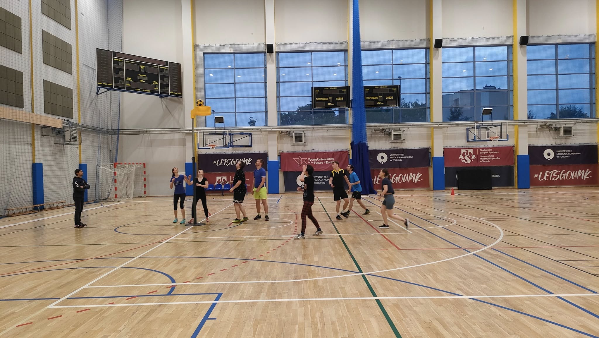 Grupa osób grających w koszykówkę na hali sportowej. Widać dwa kosze, tablicę wyników, linie boiska oraz banery promocyjne na ścianach. Przez duże okna wpada naturalne światło.